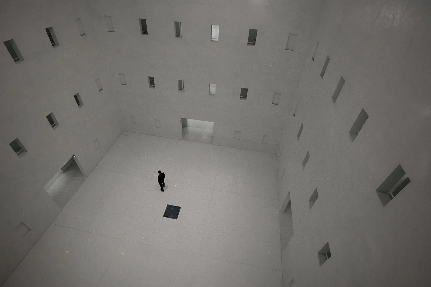 Person standing alone in a large, minimalistic white atrium with tall walls lined with small rectangular openings