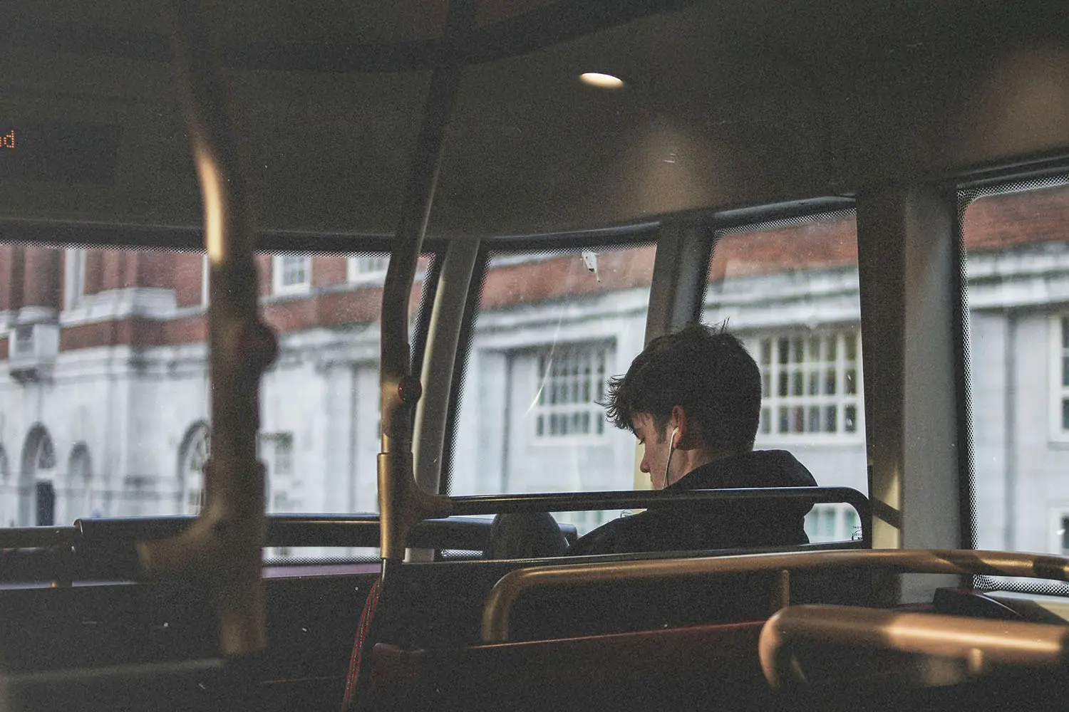 Person seated on the upper level of a bus, looking out large windows at a row of brick and white buildings outside