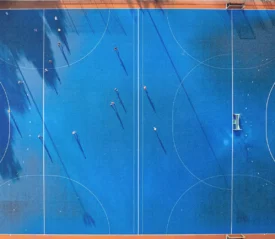 Aerial view of a bright blue sports field with players scattered across the court, casting long shadows in the sunlight