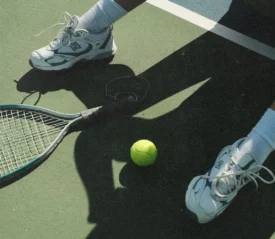 A tennis racket and bright yellow tennis ball rest on a court beside a person wearing athletic shoes and white socks