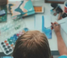Overhead view of a person painting with watercolors at a cluttered art desk