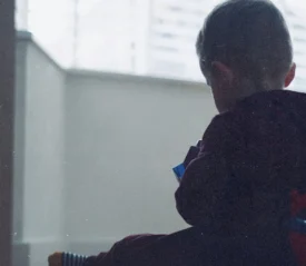 A young child sits indoors near a window, holding a toy while soft daylight filters through blinds behind them