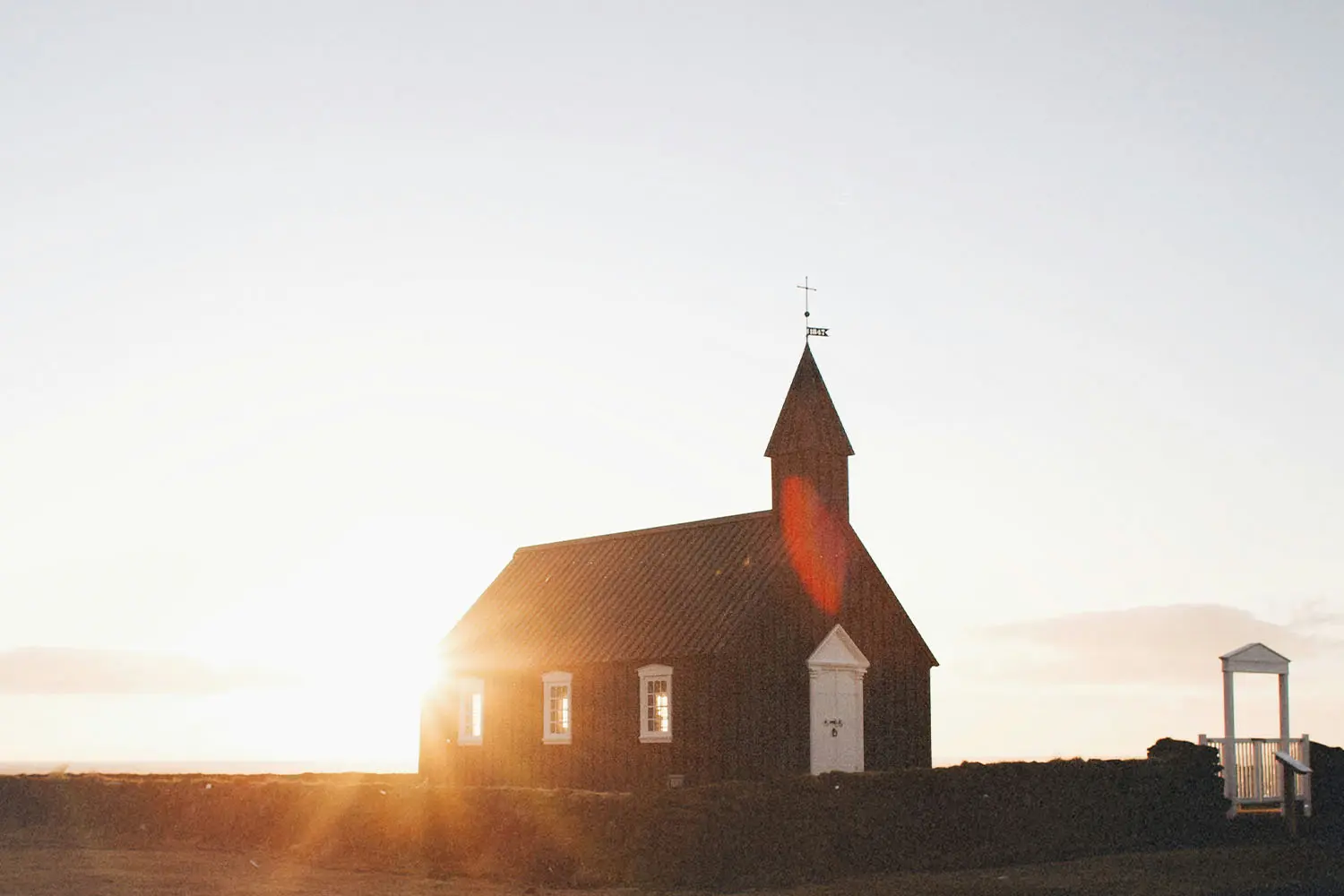 Small dark wooden church standing in an open landscape at sunrise, with warm sunlight shining behind the building and a clear sky above