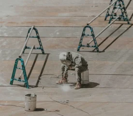 A construction worker wearing protective gear crouches on a large metal surface while using a tool that emits sparks or dust