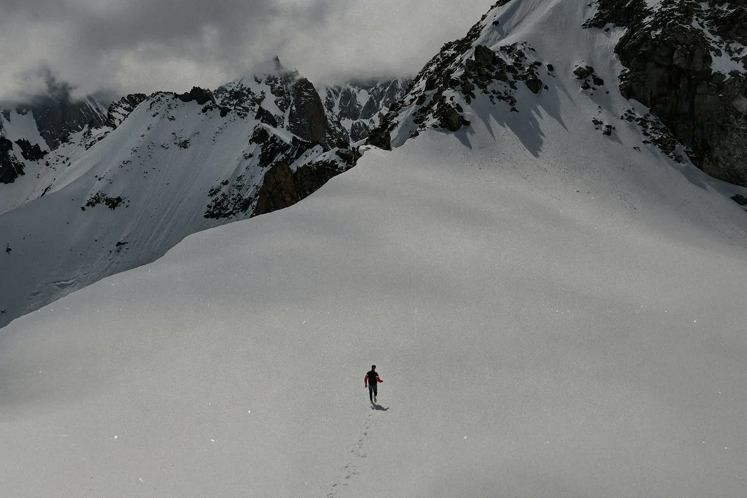 A lone person walks across a wide snowy slope surrounded by towering, rugged mountains and low-hanging clouds