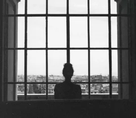 Silhouetted figure stands before a large grid‑paneled window, looking out over a sprawling cityscape