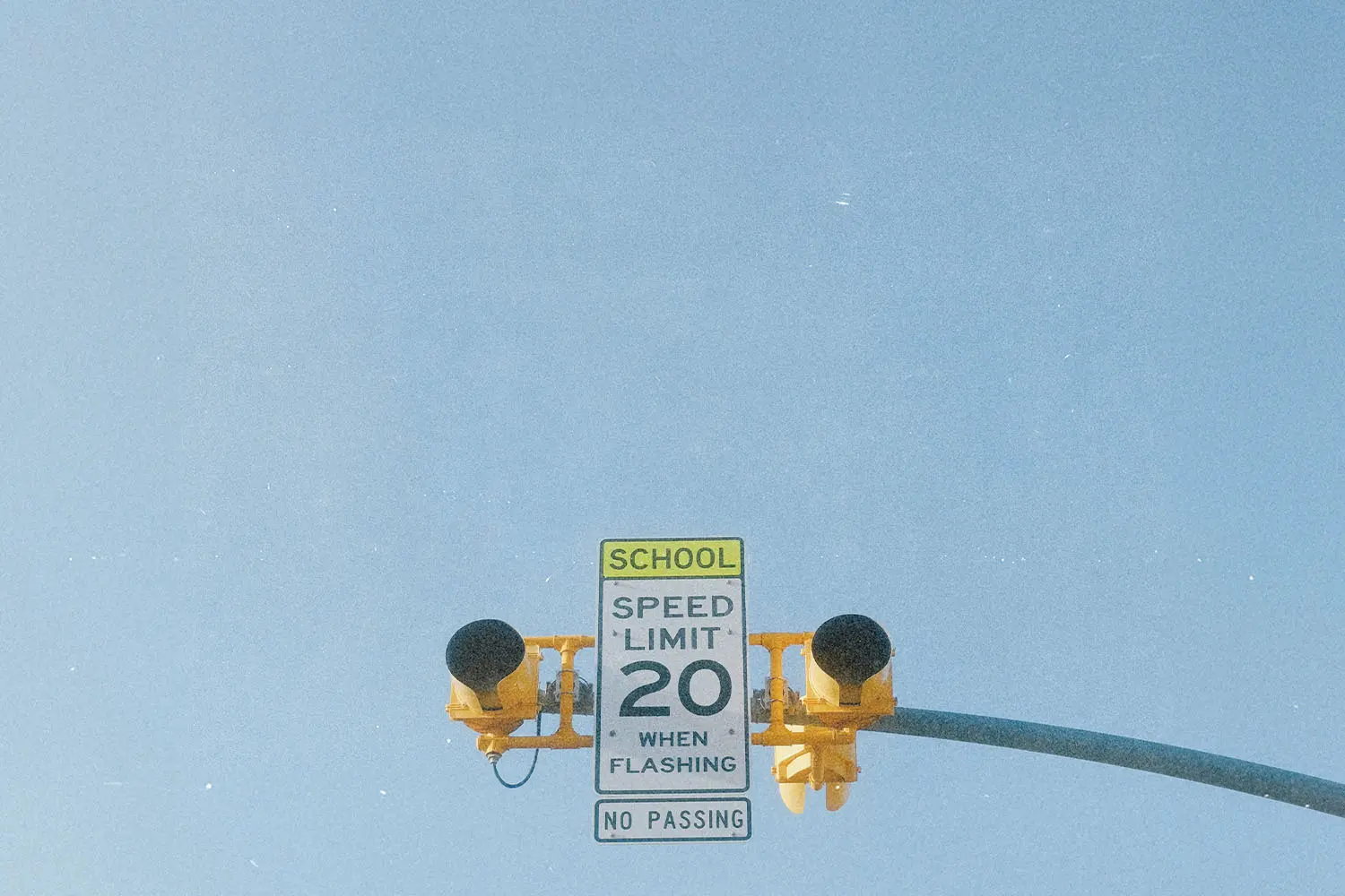School zone speed‑limit sign reading “20 when flashing,” mounted beneath yellow traffic lights against a clear blue sky