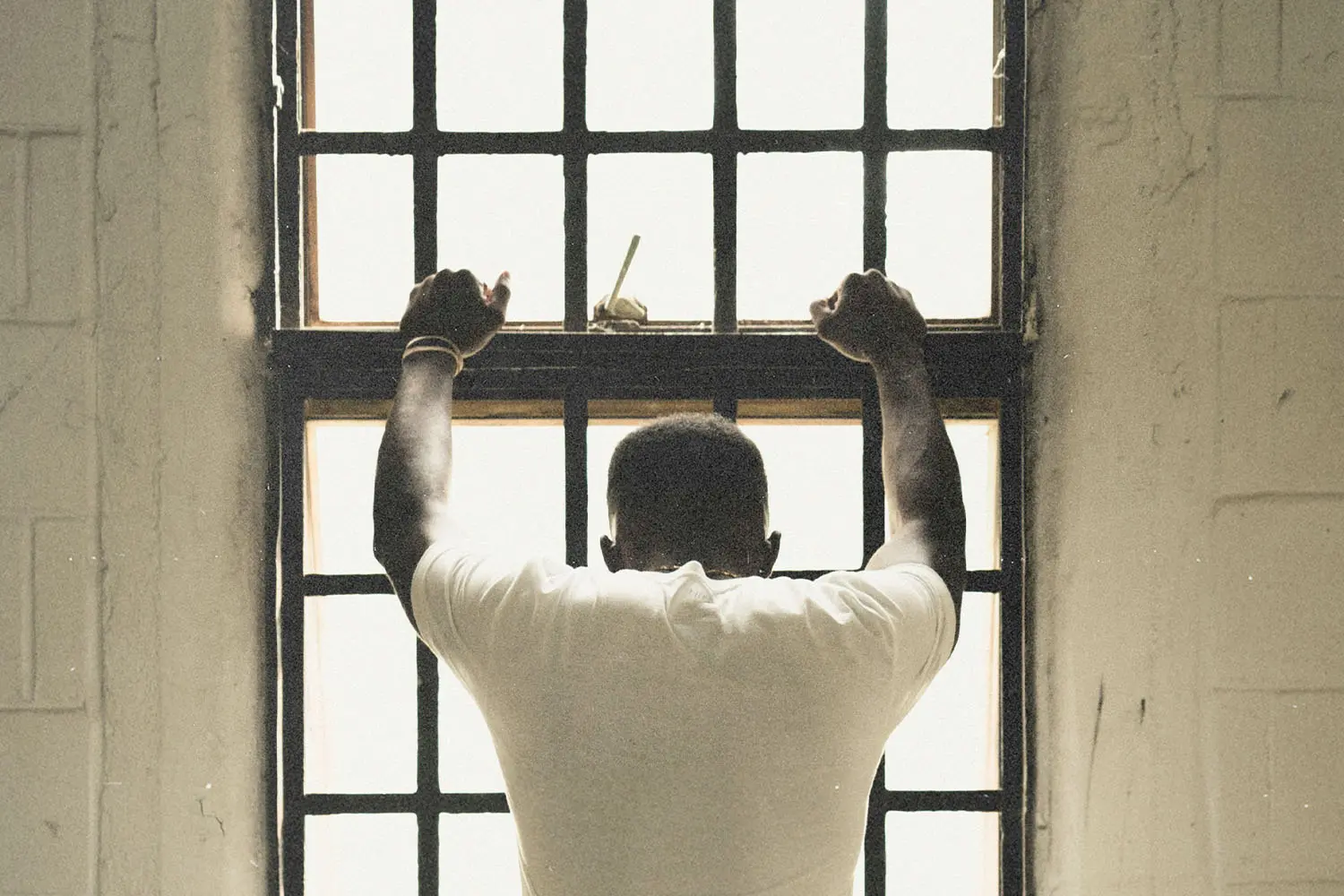 Person standing with hands gripping window bars while looking out through a bright, backlit window
