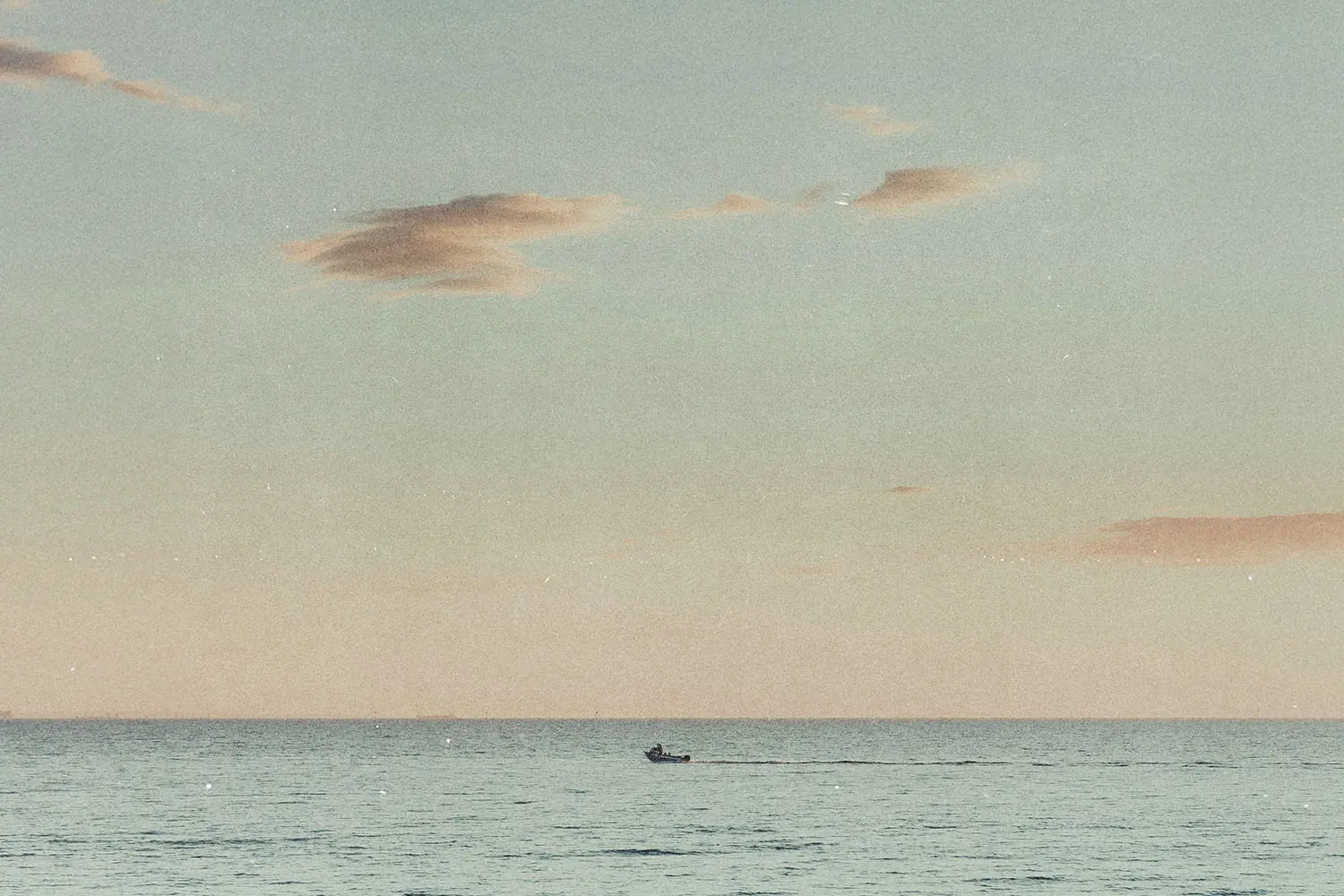 Small boat moving across a calm ocean at sunset beneath a pastel sky with scattered clouds