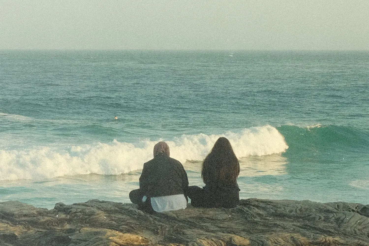 Two people sitting on rocks overlooking ocean waves rolling toward the shore