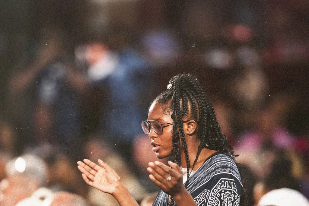 Person standing in a crowded gathering with hands raised in a worship or prayer posture, surrounded by soft, colorful lights and a blurred audience background