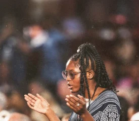 Person standing in a crowded gathering with hands raised in a worship or prayer posture, surrounded by soft, colorful lights and a blurred audience background
