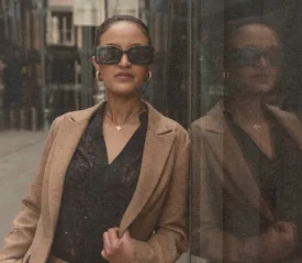 A person wearing a tan coat leans casually against a reflective glass wall in an urban setting