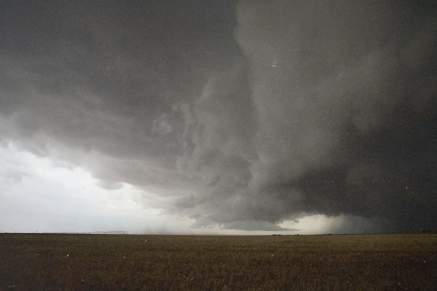 Massive storm cloud spreads dramatically across the sky above a flat, open field