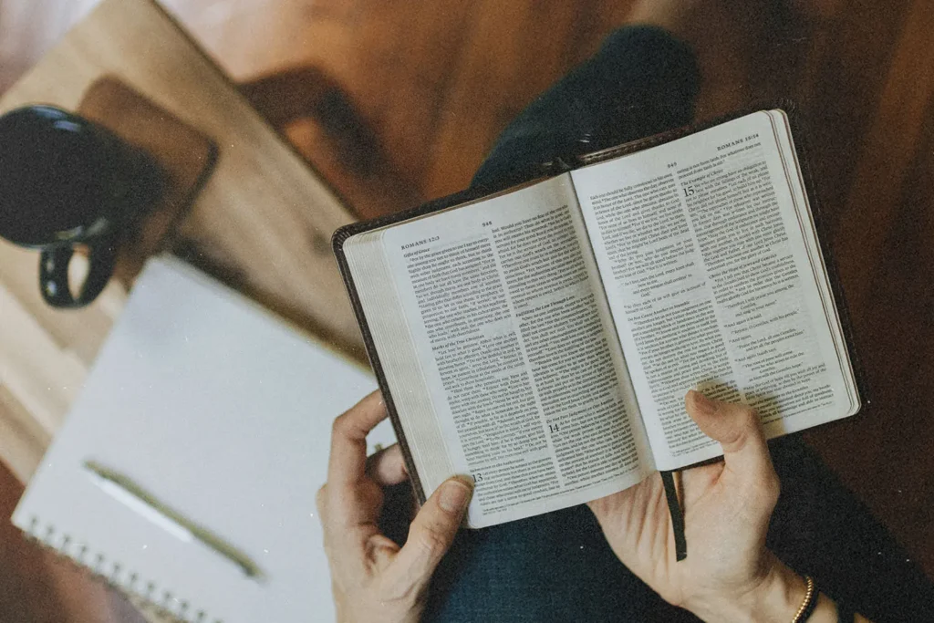 Hands hold an open Bible across the lap, with a notebook and a mug of coffee on a small table nearby