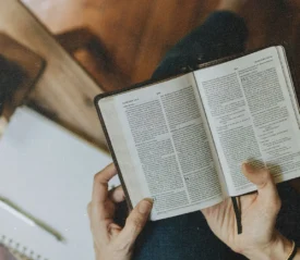 Hands hold an open Bible across the lap, with a notebook and a mug of coffee on a small table nearby
