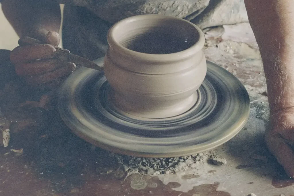 Hands shaping a clay pot on a spinning pottery wheel, with tools and wet clay visible in the workspace