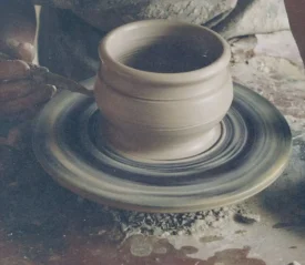 Hands shaping a clay pot on a spinning pottery wheel, with tools and wet clay visible in the workspace