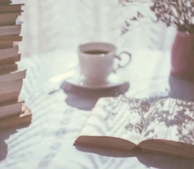 An open book rests on a sunlit table beside a tall stack of books, a cup of coffee, and a vase of flowers, with soft shadows cast across the scene