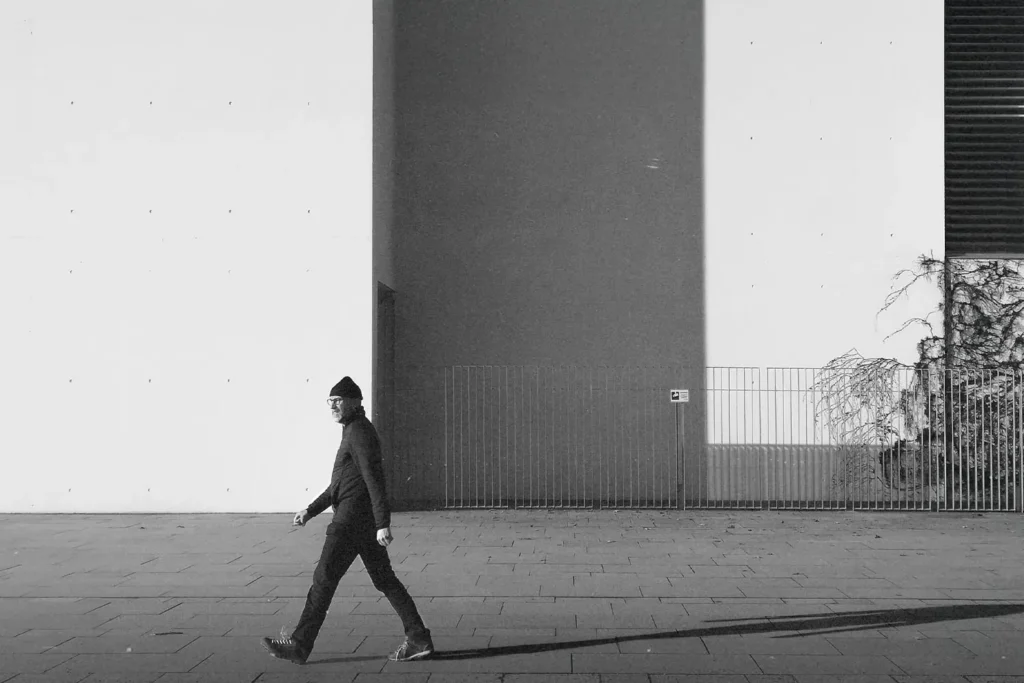 man dressed in black with glasses and a beanie hat walking down the street