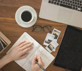 worktable with laptop, ipad, books and pictures, coffee, glasses, and a person writing in a notebook
