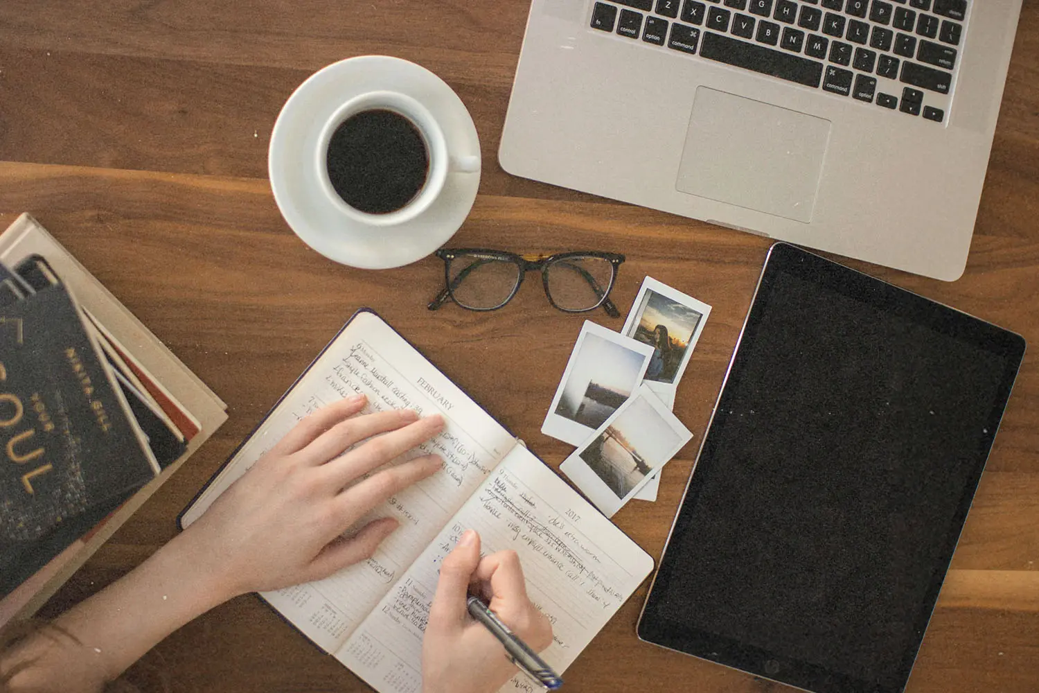 worktable with laptop, ipad, books and pictures, coffee, glasses, and a person writing in a notebook