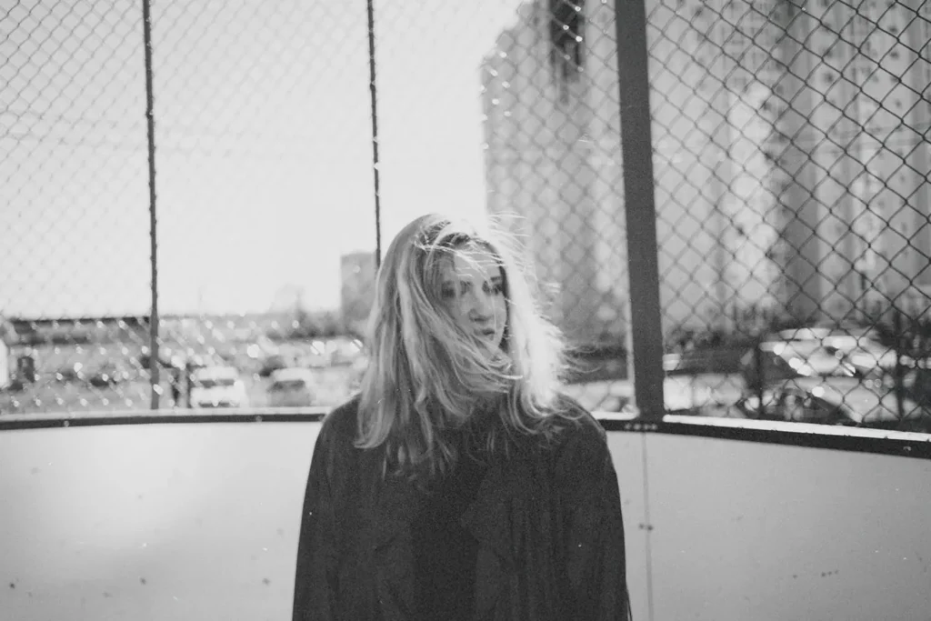 A person with long hair stands outdoors in front of a chain‑link fence, with sunlight shining through the background