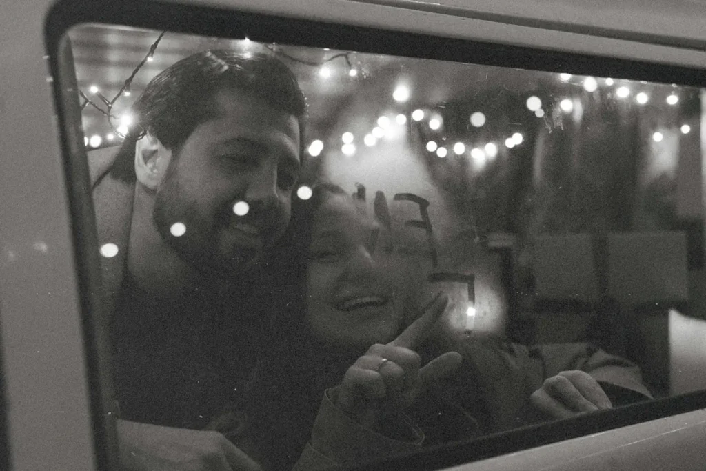 Two people look through a vehicle window adorned with string lights, writing letters in fog