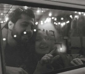 Two people look through a vehicle window adorned with string lights, writing letters in fog