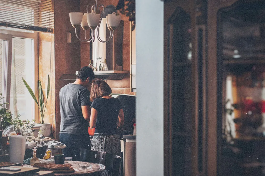 A couple standing together in a cozy kitchen preparing food at the counter, surrounded by warm lighting and houseplants