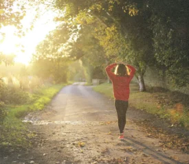 Person walking down a sunlit tree‑lined path at sunrise, wearing a red sweater and stretching with arms raised overhead