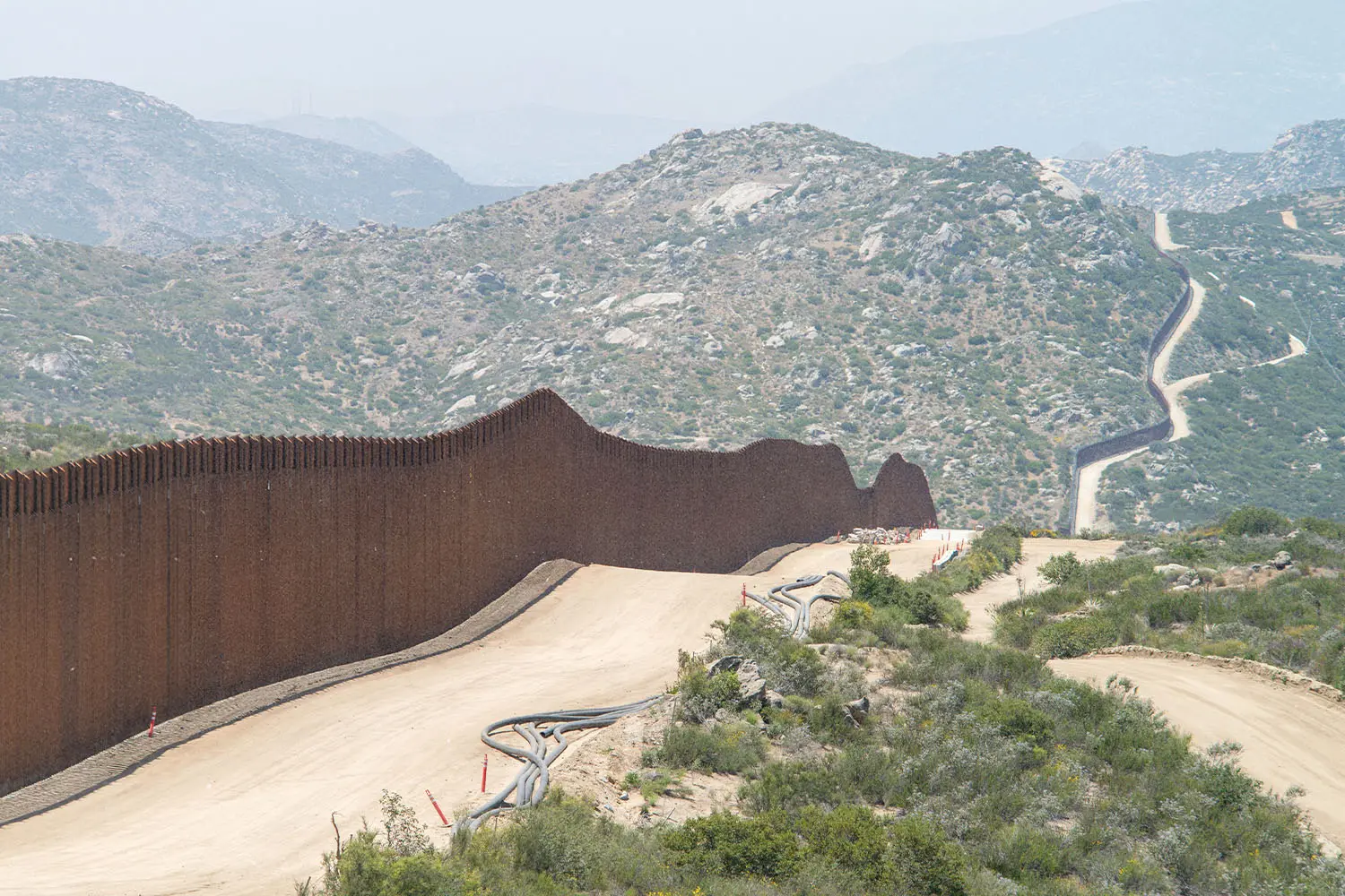 Long winding security wall running through a rugged, mountainous landscape with dirt roads following its path