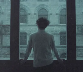 A person stands at a large window, looking out at an ornate stone building as snow falls outside