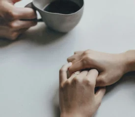 One hand holding a cup of coffee with another person sitting across the table with folded hands