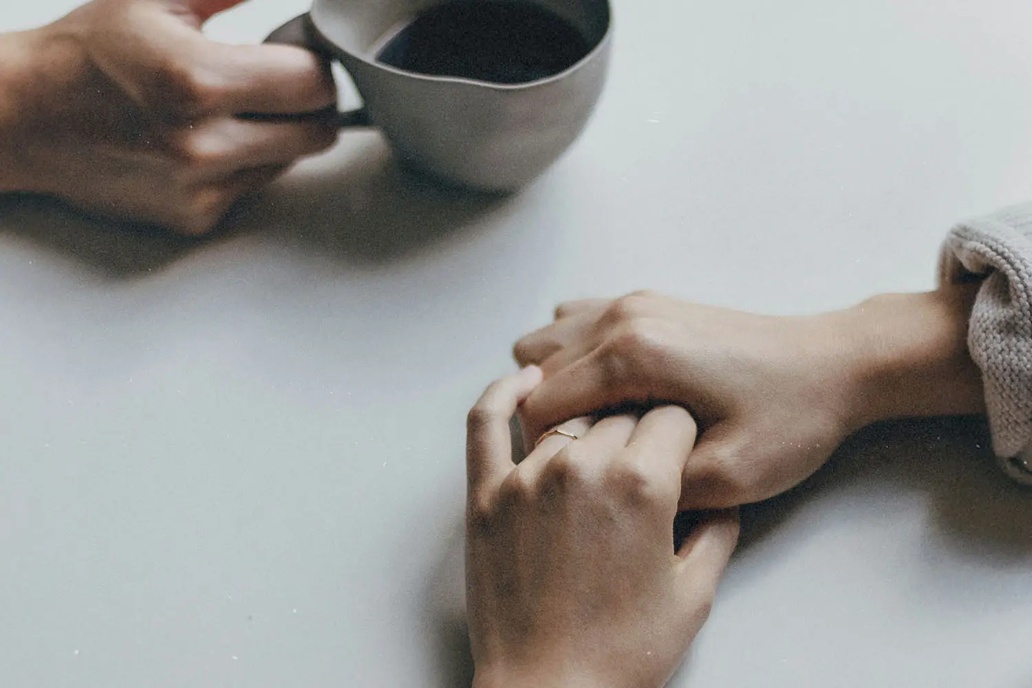 One hand holding a cup of coffee with another person sitting across the table with folded hands