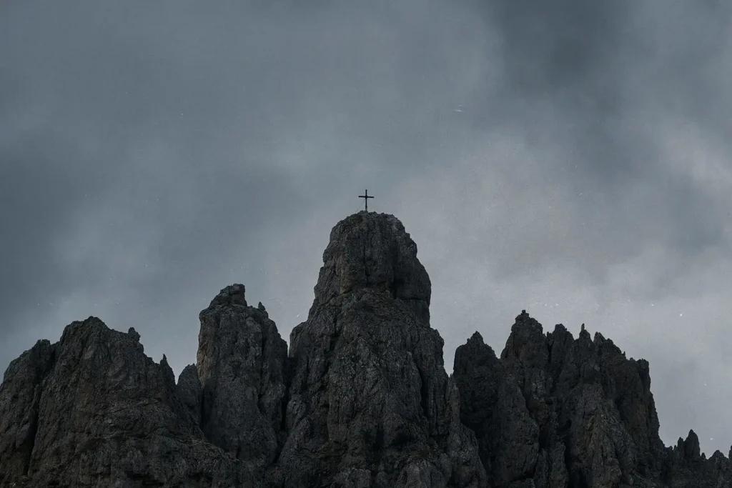 A cross stands atop a rugged mountain peak under a dark, overcast sky
