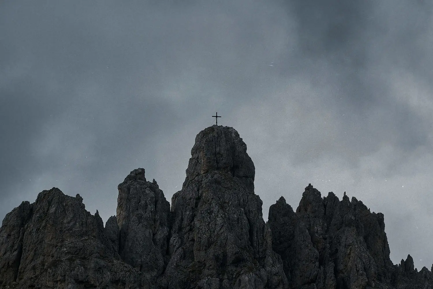 A cross stands atop a rugged mountain peak under a dark, overcast sky