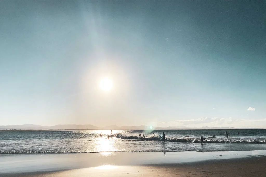 Bright sun above coastal waves with silhouettes of people in the water near the beach