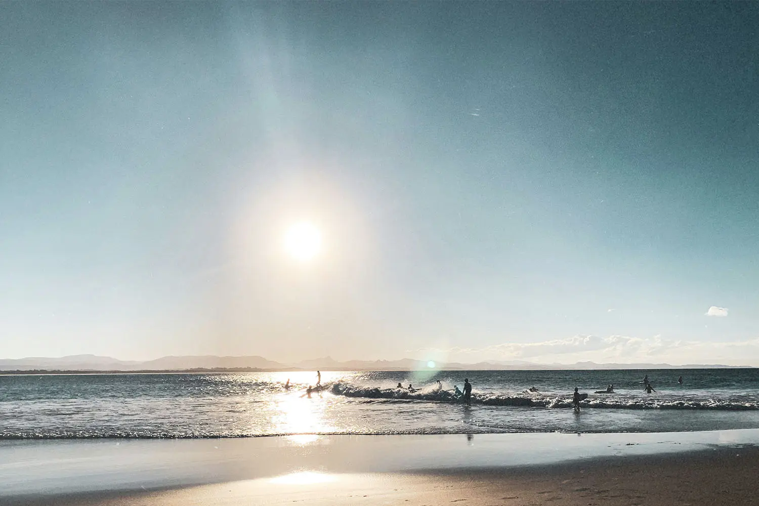 Bright sun above coastal waves with silhouettes of people in the water near the beach