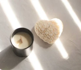 A black candle jar with a wooden wick sits beside a textured heart‑shaped object, both illuminated by bright streaks of sunlight