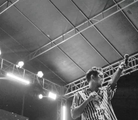 Performer on an outdoor stage holding a microphone and raising one arm under bright concert lights