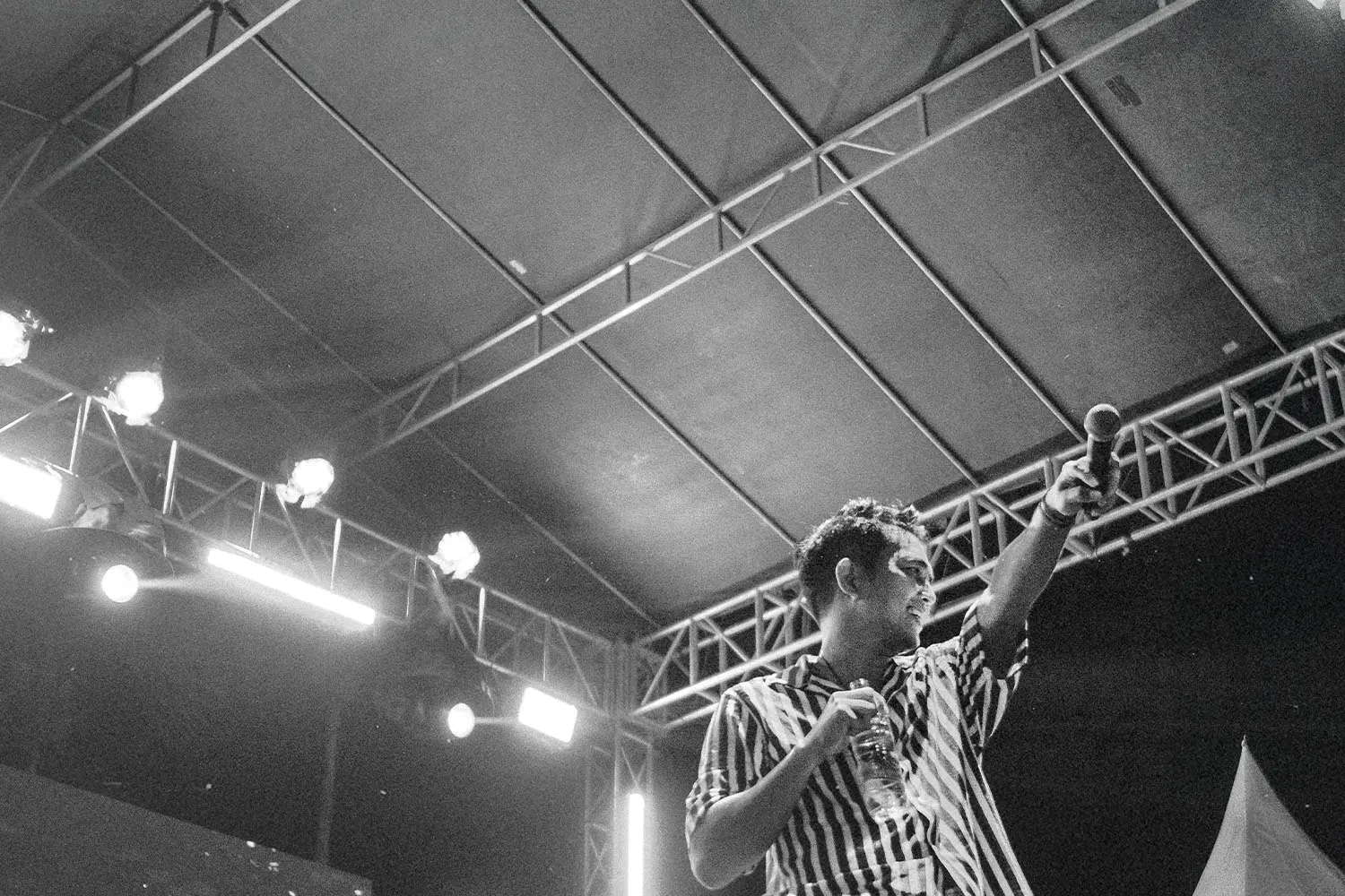Performer on an outdoor stage holding a microphone and raising one arm under bright concert lights