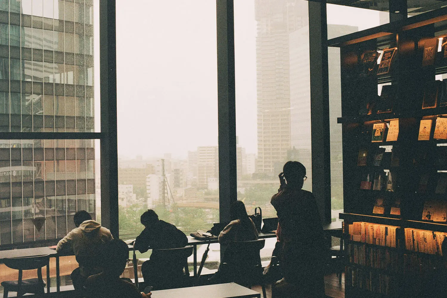 Several people sit and study near large floor‑to‑ceiling windows, overlooking a hazy cityscape from inside a modern library or café