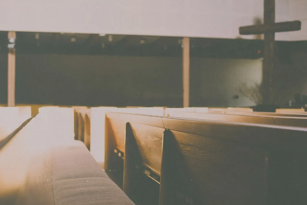 Sunlight streaming across empty wooden church pews with a large wooden cross at the front of the sanctuary