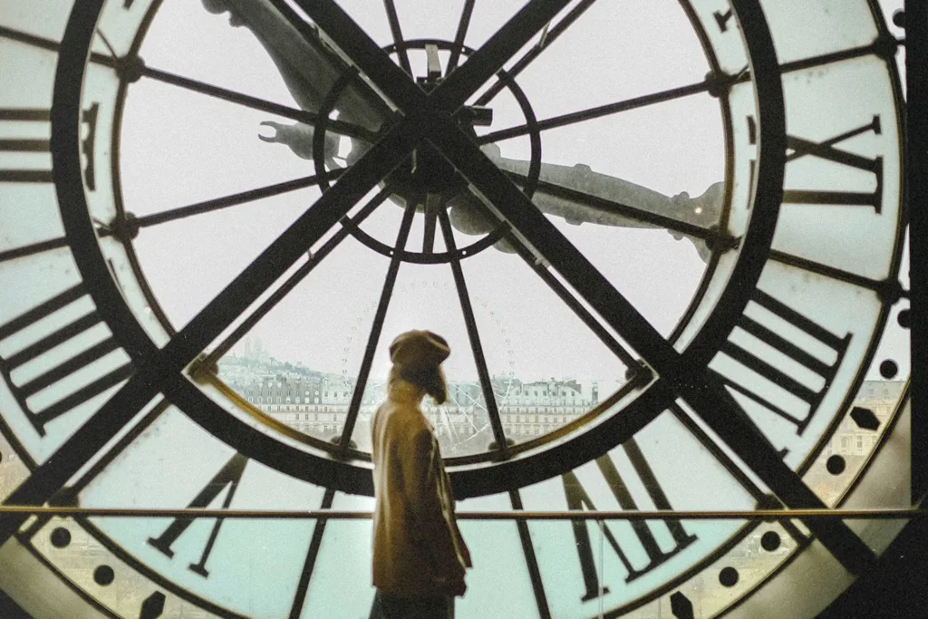 Person stands silhouetted inside a large clock tower, looking out through the transparent clock face toward a distant cityscape
