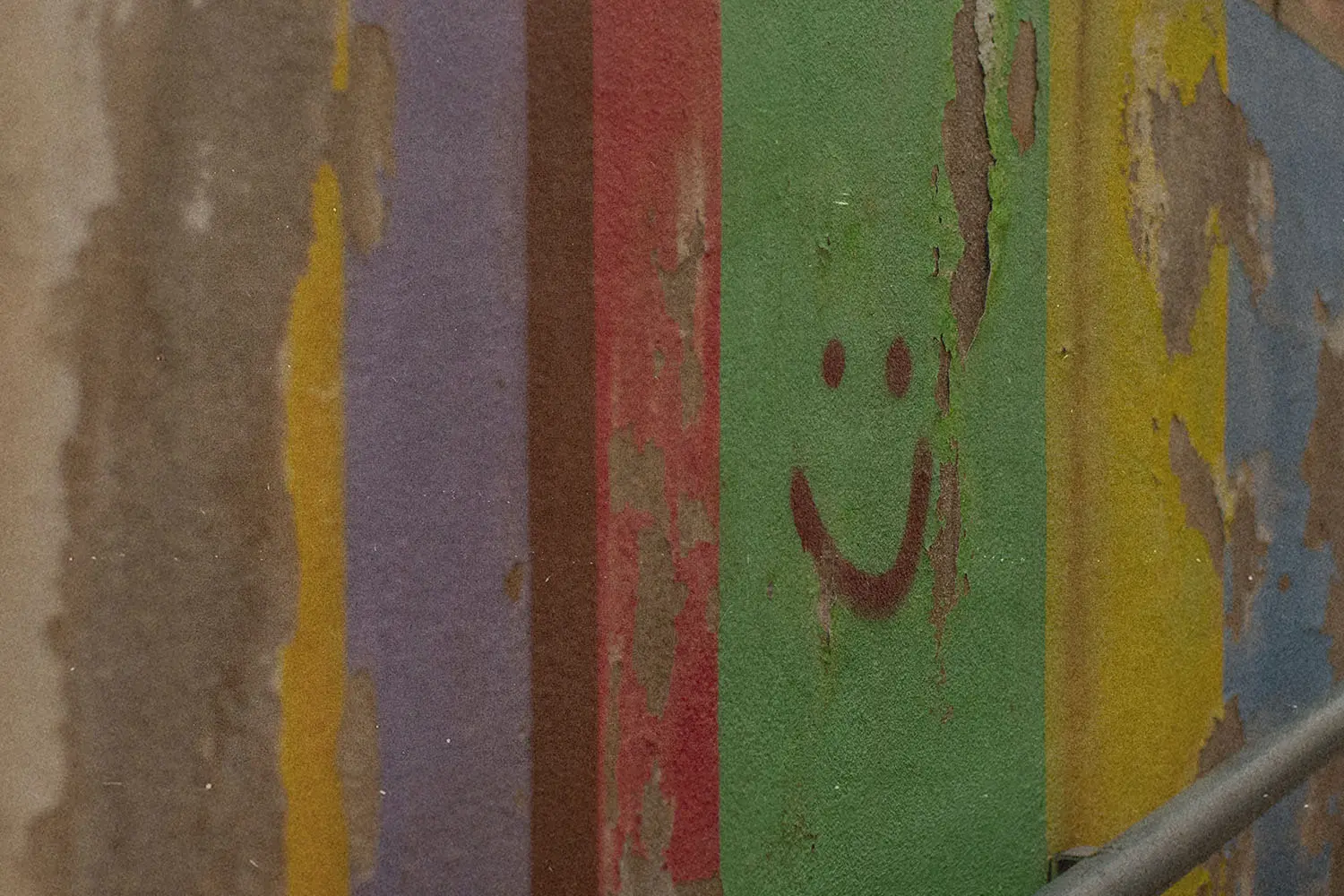 Peeling painted wall with vertical stripes of faded colors, including green, red, purple, yellow, and blue, featuring a simple smiley face drawn on the green section