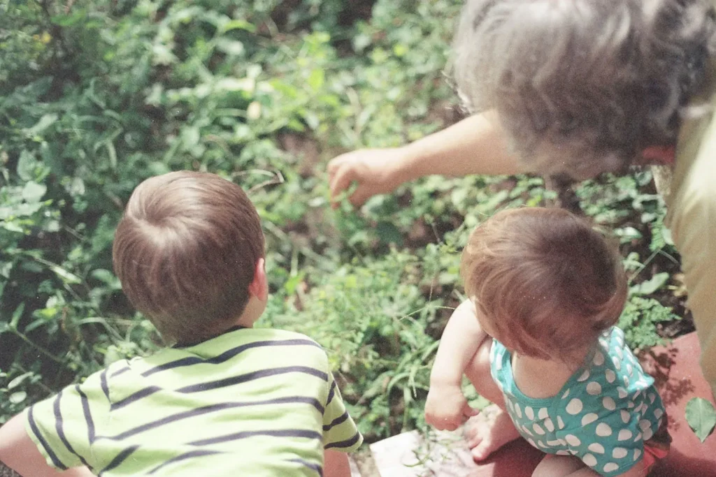 An adult and two young children lean over a garden bed while the adult points toward something in the plants