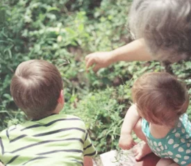An adult and two young children lean over a garden bed while the adult points toward something in the plants