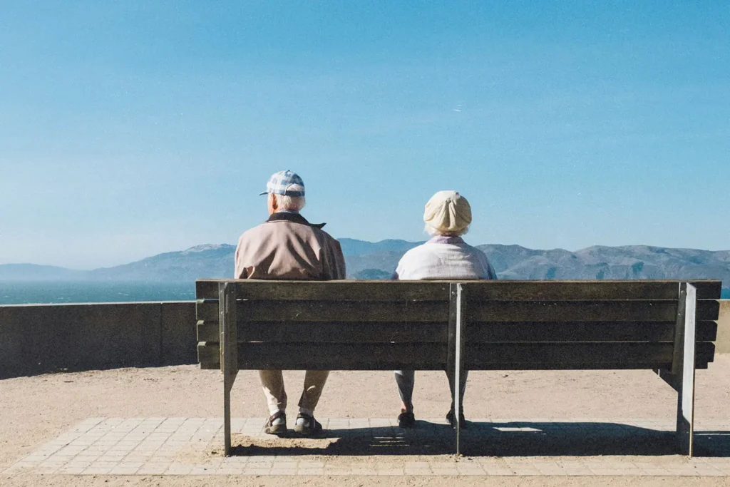Two people sit on a wooden bench overlooking a bright blue sky and distant mountains