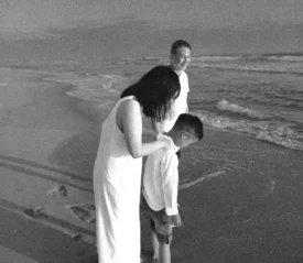 A family standing at the shoreline during sunset, with an adult gently comforting a child as waves reach their feet